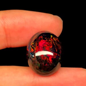 Könnte beinhalten: Ein runder, polierter schwarzer Opal-Edelstein mit einer dunklen Grundfarbe und lebhaften, intensiven roten, orangen und gelben Farbspielen auf der gesamten Oberfläche. Der Stein hat einen glatten, gewölbten Cabochon-Schliff mit einer Hochglanzoberfläche.