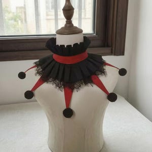 May include: A black and red ruffled collar featuring a pleated fabric design with black lace trim along the edges. The collar includes five red fabric points extending outward, each finished with a small black pom-pom at the tip of the point.