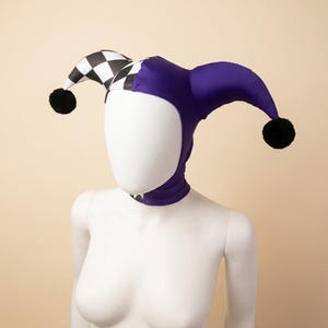 May include: A jester hat featuring a two-toned design with one side in solid purple and the other side in a black and white chequered pattern. The hat includes two pointed horns, each tipped with a small black pom-pom, and fits snugly around the head.