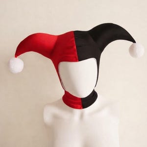 Puede incluir: Un gorro de bufón con un diseño bicolor, con un lado en rojo vibrante y el otro en negro sólido. El gorro tiene dos puntas largas y curvas que se extienden desde los lados, cada una terminada con un pompón blanco suave en el extremo.