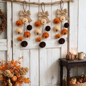 May include: Three hanging decorations suspended from a horizontal wooden branch, each featuring a hessian bow and a cluster of small fabric pumpkins in shades of orange, cream, and dark brown, arranged in a cascading vertical display.