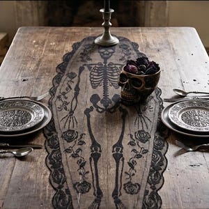 May include: A black lace table runner featuring a skeleton and rose pattern, placed on a wooden table with two plates, silverware, a skull-shaped bowl filled with dark flowers, and a metal candlestick holder at the top centre.