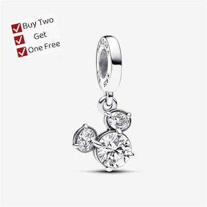 May include: A silver dangle charm featuring three round clear stones arranged in a cluster, with the text PANDORA S925 engraved on the bail. A white square graphic in the corner displays the text Buy Two Get One Free with three red checkmarks.