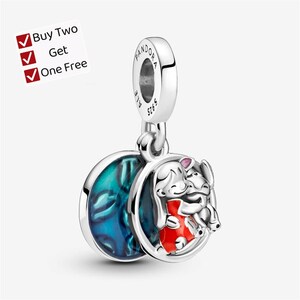 May include: A silver charm featuring a circular pendant with a blue enamel background and a raised figure of a character in a red dress hugging a small creature. The bail is engraved with Pandora and S925. Text reads: Buy Two Get One Free.