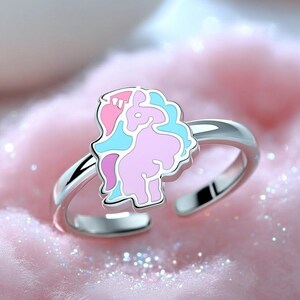 Puede incluir: Un anillo de banda abierta en tono plateado con un elemento decorativo central en forma de poni estilizado. El diseño del poni está relleno de esmalte en tonos rosa claro, rosa brillante y azul claro, creando un punto focal colorido y único.