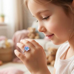 Puede incluir: Un anillo de tono plateado con un diseño de flor de cinco pétalos. Los pétalos presentan una transición de color degradado desde el rosa en los bordes exteriores hasta el azul hacia el centro, acentuado con una pequeña piedra circular azul oscuro.