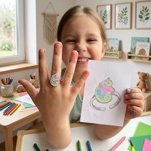 Op de afbeelding: Een zilverkleurige ring met een gestileerd eenhoornkopontwerp met pastelroze, blauwe en lichtgroene emaille accenten. De ring wordt getoond naast een witte kaart met een handgetekende illustratie van hetzelfde eenhoornringontwerp.