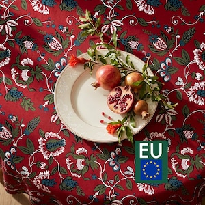 May include: A round white ceramic plate sits on a red tablecloth featuring a repeating floral pattern in green, blue, and white. The plate holds three whole pomegranates and one halved pomegranate with visible red seeds, accompanied by green leaves.