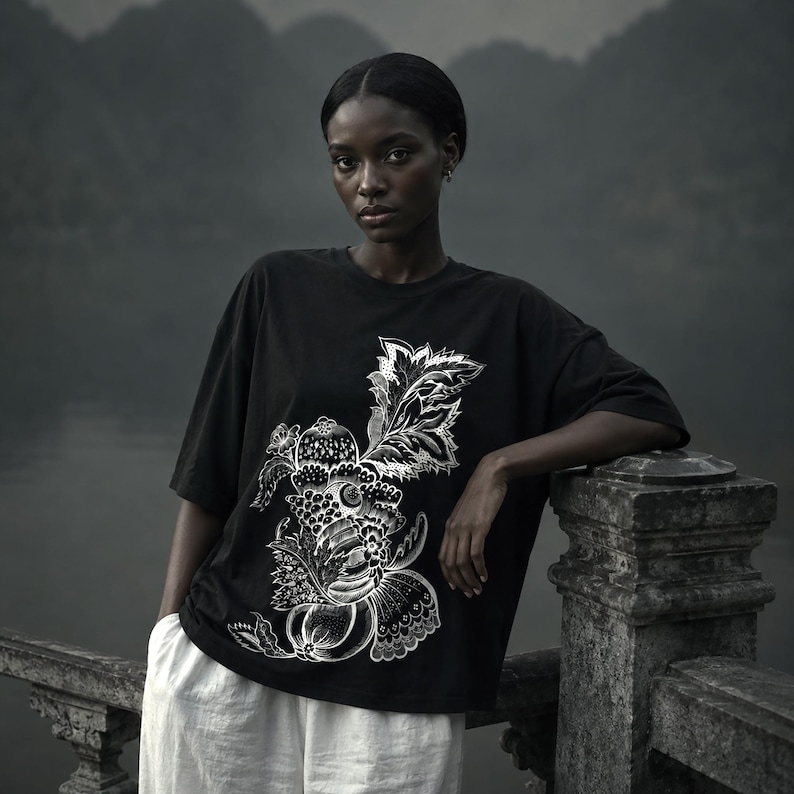 Pu&ograve; includere: Una t-shirt nera a maniche corte con una dettagliata stampa grafica bianca di elementi botanici stilizzati, tra cui foglie, frutti e motivi floreali. La maglietta presenta un girocollo e una vestibilit&agrave; ampia, indossata con pantaloni bianchi.