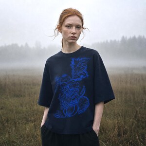 Pu&ograve; includere: Una t-shirt a maniche corte blu navy scuro con una stampa grafica prominente e intricata in un vibrante blu elettrico. Il design raffigura un insieme di elementi botanici, tra cui foglie e frutti, centrato sulla parte anteriore del capo.
