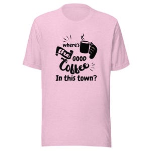 Może przedstawiać: Jasnoróżowy T-shirt z krótkim rękawem z czarnym nadrukiem graficznym przedstawiającym dłoń trzymającą kubek kawy. Tekst brzmi "where's the GOOD Coffee In this town?". Wzór znajduje się na środku klatki piersiowej melanżowej koszulki.