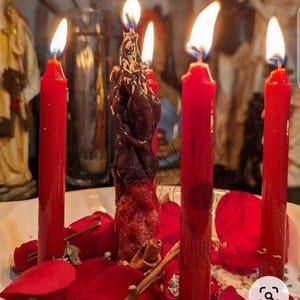 May include: A central dark brown figural candle shaped like two intertwined figures, surrounded by four tall red taper candles. The arrangement is set within a white bowl filled with scattered red rose petals and small cinnamon sticks.