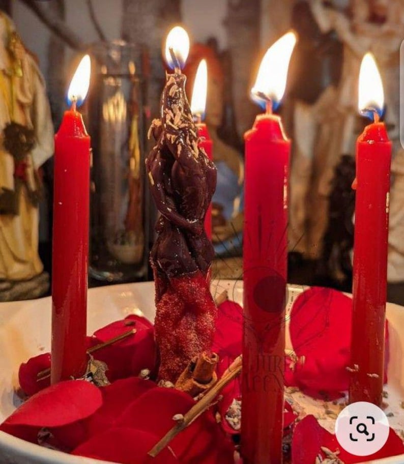 May include: A central dark brown figural candle shaped like two intertwined figures, surrounded by four tall red taper candles. The arrangement is set within a white bowl filled with scattered red rose petals and small cinnamon sticks.