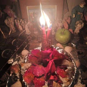 May include: Two red candles with lit flames are tied together with a red ribbon and placed in the center of a circular foil dish filled with scattered red rose petals. The arrangement is surrounded by various small decorative figurines.