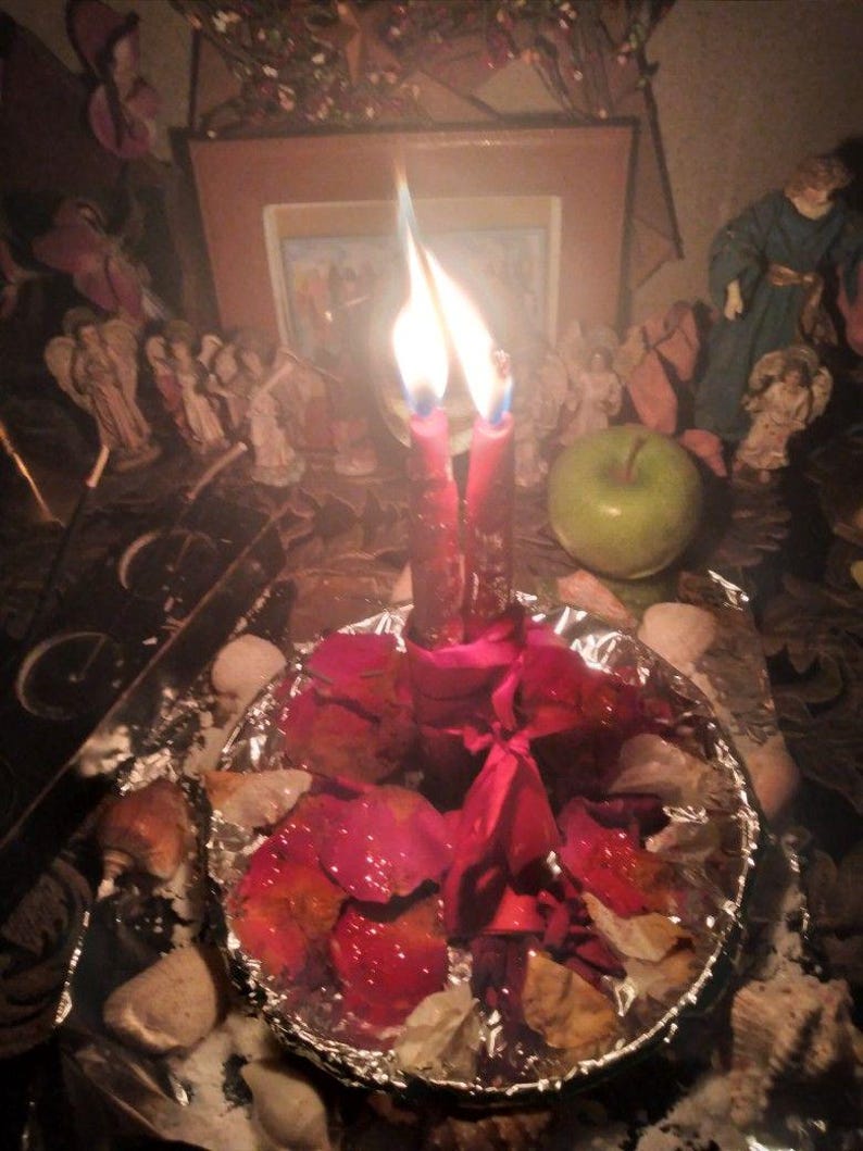May include: Two red candles with lit flames are tied together with a red ribbon and placed in the center of a circular foil dish filled with scattered red rose petals. The arrangement is surrounded by various small decorative figurines.