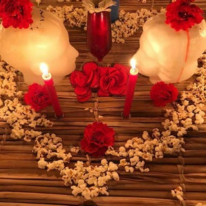 May include: A decorative arrangement featuring two white head-shaped candles topped with red flowers, a central red glass vase with a white flower, a blue pillar candle, two red taper candles, and scattered popcorn pieces on a brown woven mat.