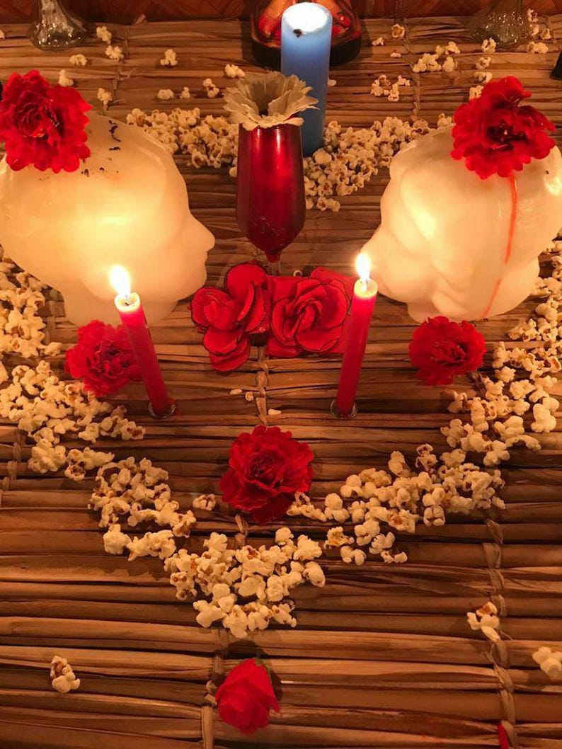 May include: A decorative arrangement featuring two white head-shaped candles topped with red flowers, a central red glass vase with a white flower, a blue pillar candle, two red taper candles, and scattered popcorn pieces on a brown woven mat.