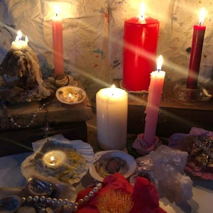 May include: A collection of lit candles in various sizes and colors, including red, pink, and white, arranged with seashells, pearl strands, and a large red flower. The scene features wax-covered surfaces and glowing flames casting light.