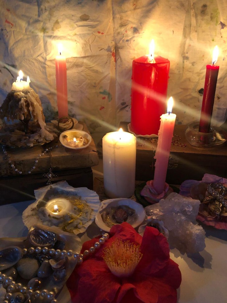 May include: A collection of lit candles in various sizes and colors, including red, pink, and white, arranged with seashells, pearl strands, and a large red flower. The scene features wax-covered surfaces and glowing flames casting light.