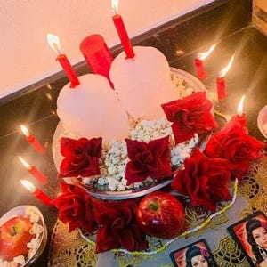 May include: A silver platter holds two white skull-shaped candles with red tapers, surrounded by popcorn and red fabric roses. A red apple sits nearby, accompanied by several small lit red candles and two rectangular cards featuring portraits.