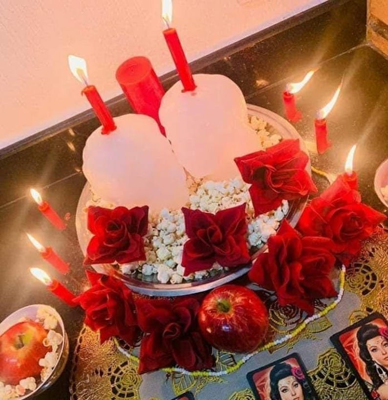 May include: A silver platter holds two white skull-shaped candles with red tapers, surrounded by popcorn and red fabric roses. A red apple sits nearby, accompanied by several small lit red candles and two rectangular cards featuring portraits.