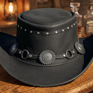 Puede incluir: Un sombrero de vaquero de cuero negro con copa plana y ala curva. El sombrero está decorado con una banda de tachuelas metálicas plateadas alrededor de la copa y una segunda banda con conchos metálicos circulares y anillos en la base.