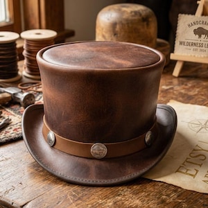 May include: A dark brown leather top hat featuring a smooth, cylindrical crown and a curved brim. A lighter brown leather band encircles the base of the crown, decorated with three silver-toned metal coins embossed with a buffalo design.