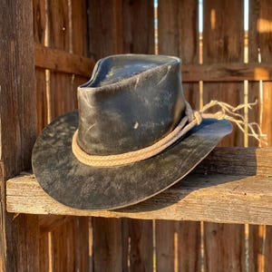 Könnte beinhalten: Ein dunkel anthrazitfarbener Cowboyhut mit einer verwitterten, abgenutzten Textur. Der Hut hat eine hohe Krone und eine breite, geschwungene Krempe. Ein dickes, hellbeiges geflochtenes Seilband umschließt die Basis der Krone und endet seitlich.