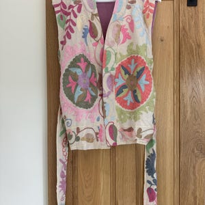 May include: A sleeveless waistcoat in a light beige fabric featuring vibrant, multi-coloured floral and circular embroidery in shades of pink, green, blue, and brown. The garment has an open front, a high collar, and long, matching embroidered tie straps.