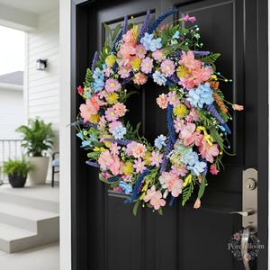 Può includere: Una ghirlanda floreale circolare caratterizzata da un mix vivace di fiori rosa, azzurri e gialli intervallati da spighe viola simili alla lavanda e fogliame verde, montata su una porta nera con il testo PorchBloom visibile vicino alla maniglia.