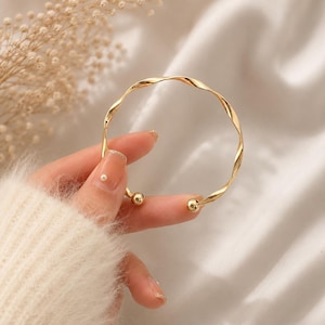 May include: A gold-toned metal cuff bracelet featuring a twisted, spiral-like band design. The bracelet is open-ended and terminates at each end with a small, smooth, polished gold-toned sphere. The metal has a reflective, high-shine finish.