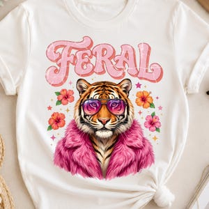 Puede incluir: Una camiseta blanca con el gráfico de un tigre que lleva gafas de sol con cristales rosas y un abrigo de piel sintética rosa. El tigre está rodeado de flores de hibisco naranjas y rosas y pequeñas estrellas. La palabra FERAL aparece en letras rosas.