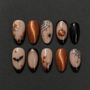 May include: A set of ten press-on nails featuring a Halloween theme with a nude base. Designs include metallic gold leaves, shimmering copper, black spiderwebs with spiders, a pumpkin in a gold frame, a black bat, and small gold stars.