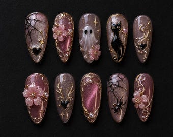 May include: A set of ten almond-shaped press-on nails featuring a mauve cat-eye base. Designs include black spiderwebs, spiders, a white ghost, a black cat, pink floral accents, gold filigree, small pearls, and black heart-shaped charms.