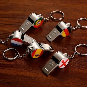 May include: Five silver-toned metal whistles attached to circular key rings. Each whistle features a circular enamel inlay displaying a different national flag, including Portugal, Germany, the Netherlands, Spain, and England, with a stamped logo.