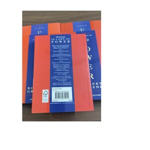 May include: A stack of paperback books titled The Concise 48 Laws of Power by Robert Greene. The back covers are orange with a central blue vertical stripe containing white text, a barcode, and an FSC logo. The books are arranged in a cluster.