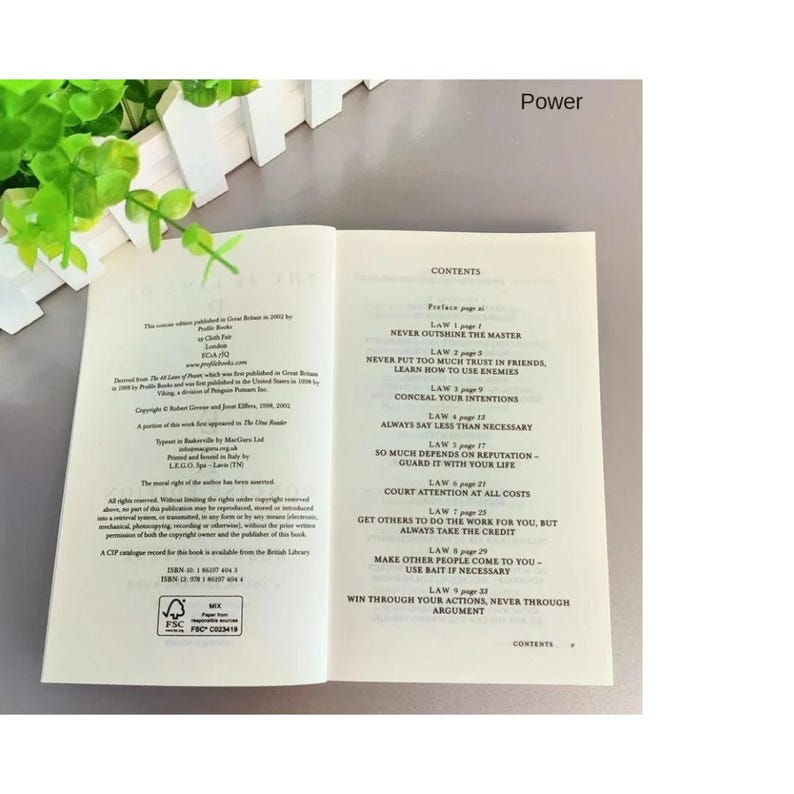 May include: An open book displaying the contents page with the title Power at the top. The left page shows publication details and copyright information, while the right page lists nine laws, including Never Outshine the Master and Conceal Your Intentions.