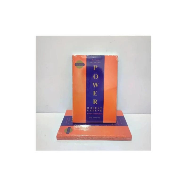 May include: A book titled The 48 Laws of Power by Robert Greene, featuring a vertical blue central stripe on an orange cover. The book is displayed in two positions: one standing upright and one lying flat underneath it, both wrapped in clear plastic.