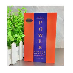 May include: The book titled The Concise 48 Laws of Power by Robert Greene features a vertical design with a central dark blue stripe flanked by orange panels. The text is printed in gold and white, including a gold circle badge labeled International Bestseller.