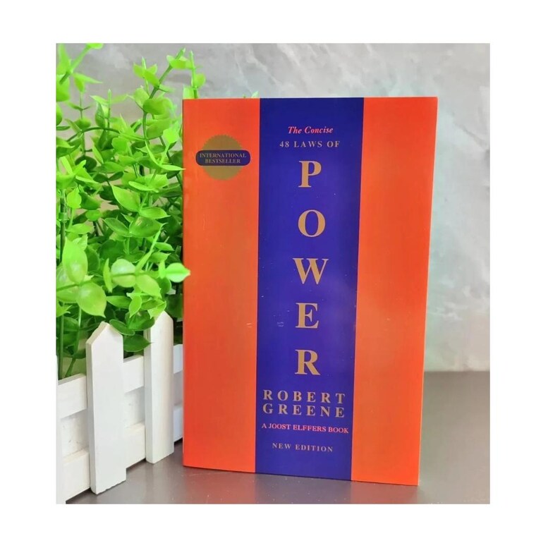 May include: The book titled The Concise 48 Laws of Power by Robert Greene features a vertical design with a central dark blue stripe flanked by orange panels. The text is printed in gold and white, including a gold circle badge labeled International Bestseller.