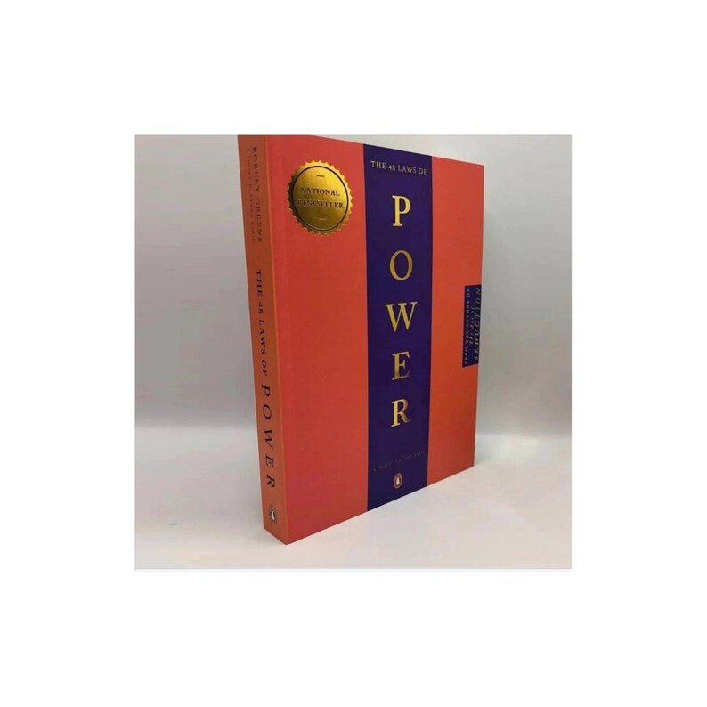 May include: A hardcover book titled The 48 Laws of Power by Robert Greene. The cover is orange with a vertical purple stripe featuring the word POWER in gold lettering. A gold circular seal is on the upper left, and a small penguin logo is at the base.
