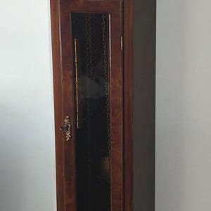 Peut inclure: Une horloge grand-père en bois foncé avec un cadran circulaire doré orné de chiffres romains et d'un indicateur de phases de lune. La porte vitrée révèle des chaînes et un pendule, le tout surmonté de trois fleurons décoratifs en laiton.