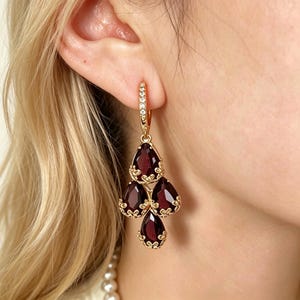 May include: A gold dangle earring featuring a hoop encrusted with small clear stones and a cluster of four deep red, pear-shaped faceted gemstones. Each gemstone is set in a decorative gold filigree frame, creating a tiered, chandelier-style drop.