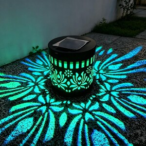 Peut inclure: Une lanterne solaire cylindrique noire avec un motif floral découpé qui projette une lumière turquoise vive sur le sol. Le dessus de la lanterne comporte un panneau solaire carré et les côtés émettent une lueur à travers le motif complexe.