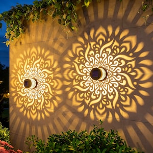 May include: Two circular solar-powered wall lights mounted on a tiled surface, each emitting a warm, bright light that projects a large, intricate, symmetrical mandala-like pattern onto the wall, creating a decorative shadow effect.