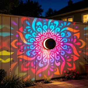 Puede incluir: Una lámpara de pared solar circular montada en una valla, que proyecta un vibrante patrón de mandala multicolor en tonos rosa, naranja y cian sobre los listones de madera verticales, creando una exhibición de luz simétrica por la noche.