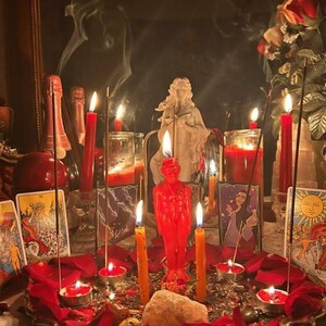 May include: A collection of red and orange candles, including a central figure-shaped candle, arranged on a tray with scattered red rose petals, incense sticks, a white stone, and tarot cards featuring illustrations of figures and the sun.