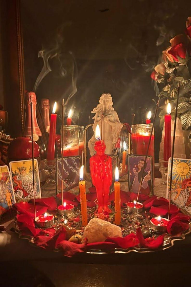 May include: A collection of red and orange candles, including a central figure-shaped candle, arranged on a tray with scattered red rose petals, incense sticks, a white stone, and tarot cards featuring illustrations of figures and the sun.