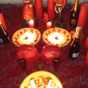 Puede incluir: Un mantel de encaje rojo sostiene dos cuencos de cer&aacute;mica llenos de fresas en rodajas y crema, un tercer cuenco con fruta, dos velas rojas altas, dos velas rojas peque&ntilde;as, dos figuras, dos copas de vino y dos botellas de champ&aacute;n.