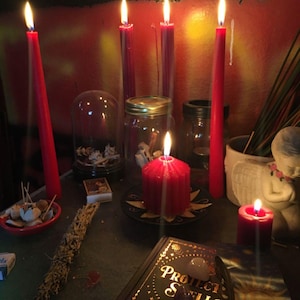May include: A collection of red candles, including tall tapers and a thick pillar candle, arranged with a book titled Protection Spells, a glass jar, a small glass dome, a white angel figurine, and a bundle of dried herbs on a dark surface.